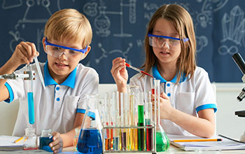 A boy and a girl pour liquid in a test tube using pipette.  Multiple test tubes on the table with colorful liquids inside